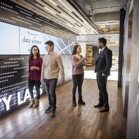 Students walking in a hallway with a huge data screen visible on the wall.